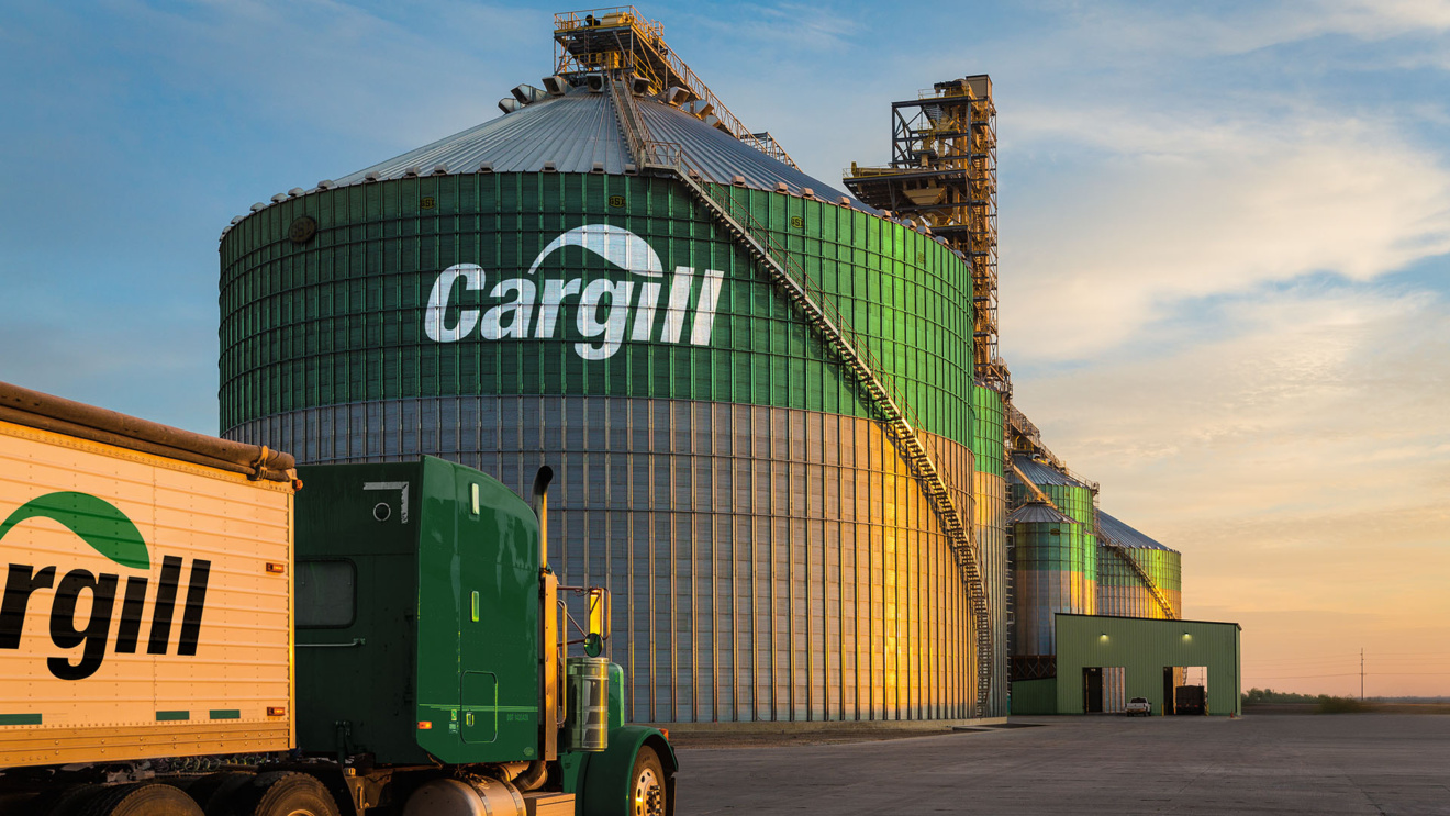 "A Cargill branded semi-truck is parked in front of large grain silos at a Cargill facility. The silos are silver and green with the Cargill logo prominently displayed."